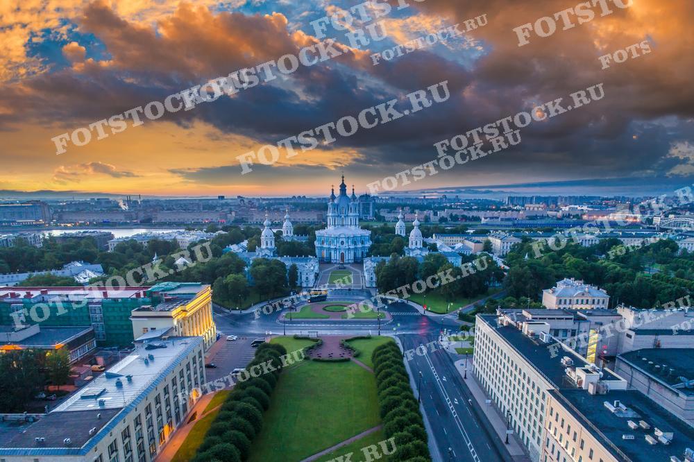 На фотографии изображен восхитительный вид на Санкт-Петербург с высоты птичьего полета во время заката. Центральным объектом является Смольный собор, его белоснежные стены и голубые купола выделяются на фоне городской застройки. Небо украшено облаками, окрашенными в теплые оттенки заката. Это изображение идеально подходит для использования в туристических проспектах, образовательных материалах и публикациях, посвященных архитектуре и истории города.